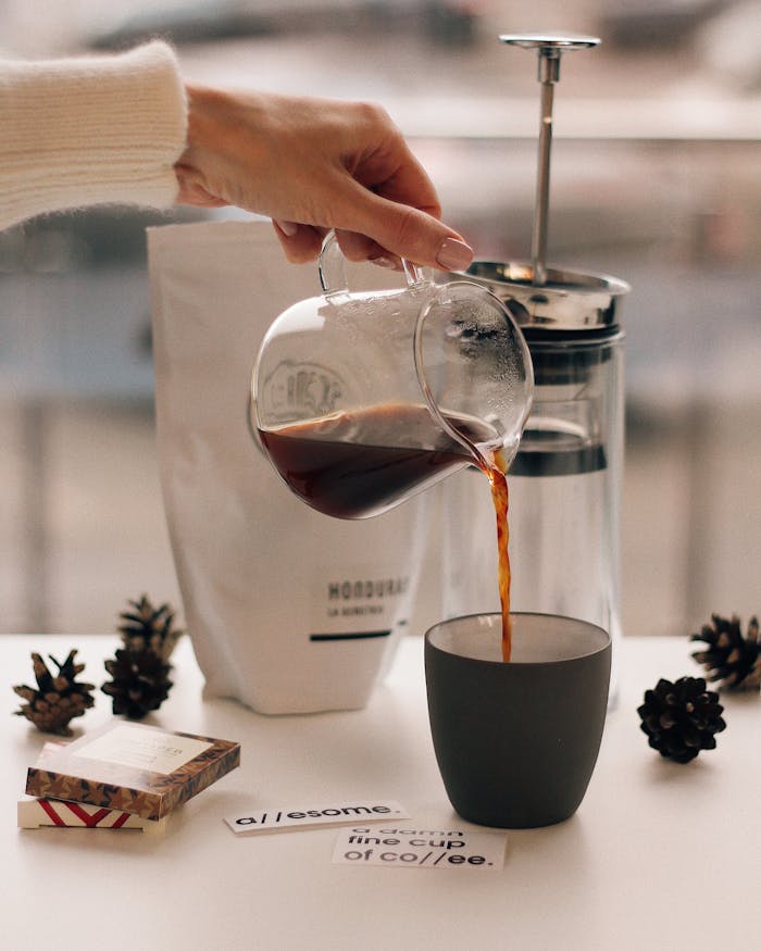 A hand pours coffee into a cup alongside a French press, creating a cozy morning vibe.